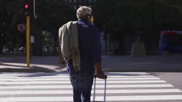 Rear view of african american senior man with trolley bag crossing the road