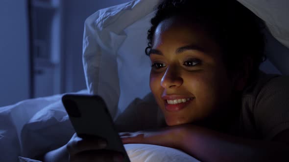 Woman with Smartphone Under Duvet in Bed at Night