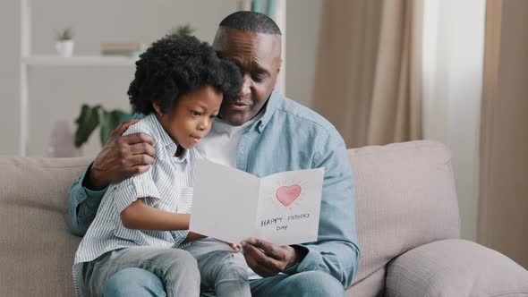 Mature African American Man with Cute Kid Girl Hugging Sitting in Room on Sofa Father Reads Greeting
