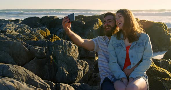 Smiling happy young couple clicking selfie on rock at the beach 4k