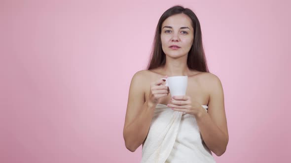 Woman in Bath Towel with White Cup Drinks Coffee Isolated on Pink Background