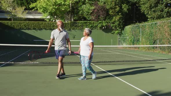 African american senior couple holding rackets talking to each other on the tennis court