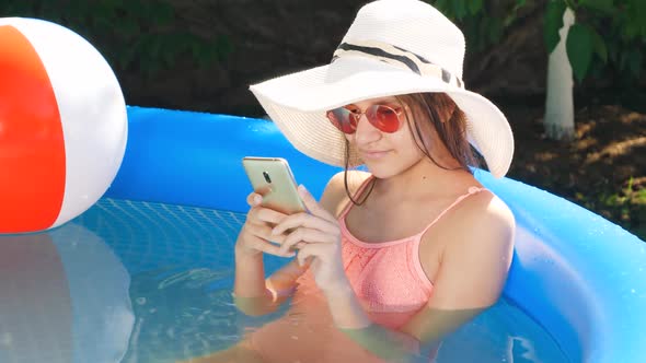 Portrait of Beautiful Young Girl Relaxing in Outdoor Swimming Pool at Backyard and Typing Message on