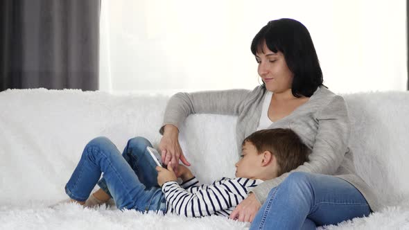 Happy Family: Mother and Son Spend Time Playing with a Tablet. Woman and Child Sitting on the Sofa