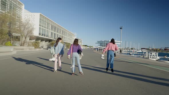 Three Girls Roller Skate on Road By Water While Holding Their Shoes