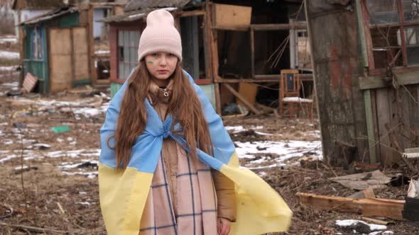 a girl wearing in the Ukrainian flag looks sadly at the camera against the background of destroyed h