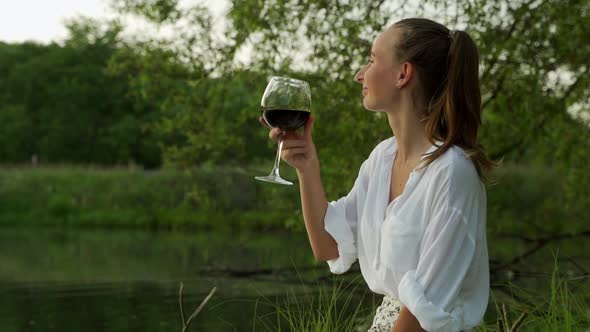 Side View Young Woman Drinks Wine From a Glass Sitting By the Lake at Sunset