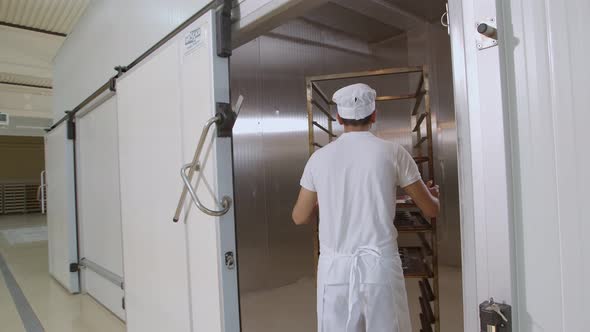 Baker Dressed in White and Toque Puts a Cart with Bread Into Big Oven