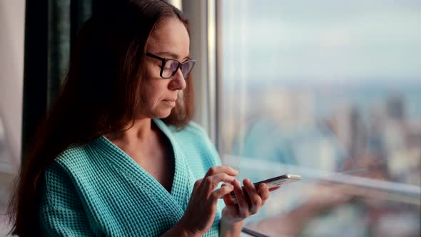 Portrait of a Woman with Glasses in the Morning in a Blue Robe Standing at the Window and Answering