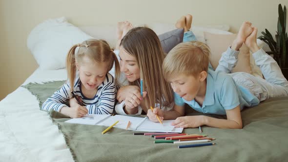 Family Leisure Mom and Kids Lay on Bed and Draw Pictures on Paper in Apartment
