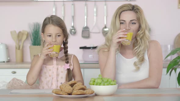 Happy Caucasian Mother and Daughter Drinking Healthful Orange Juice, Talking, and Smiling at Camera