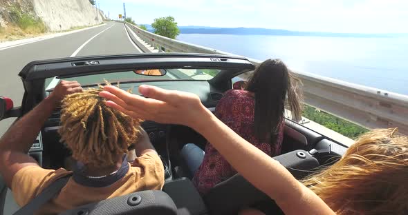 Friends dancing and waving arms driving on road along coast in red convertible