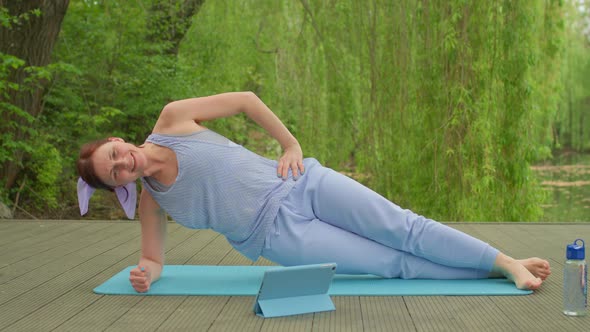 30s Woman in Sportswear Doing Aerobics Plank Exercise Using Tablet Computer Outdoors