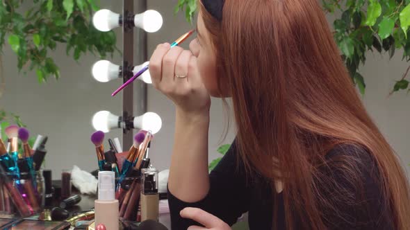 Young Woman Applying Makeup While Sitting at Her Vanity Table with Lots of Cosmetics