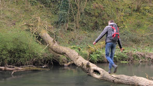 Young Man with Backpack Crossing the Mountain River Along a Fallen Tree 