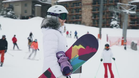 Woman with Snowboard Walking By Snowy Mountain with Skiers Riding Down Slope