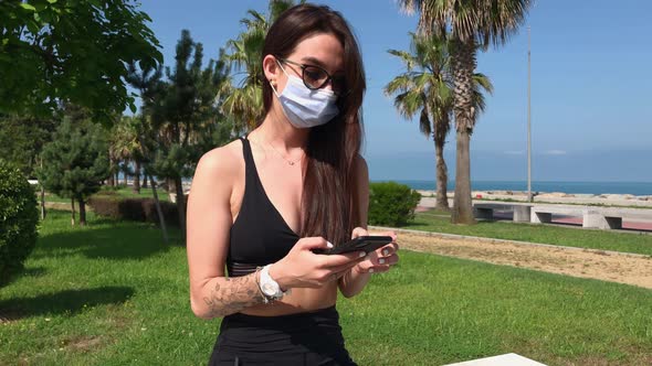 Girl Wearing Face Medical Mask Uses Smartphone in the Park.