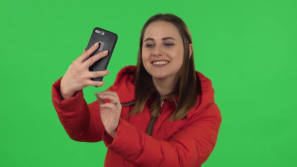 Portrait of Lovely Girl in a Red Down Jacket Is Making Selfie on Mobile Phone Then Looking Photos