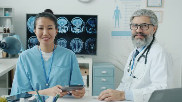 Doctor and Nurse Sitting in Workplace in Hospital Smiling and Looking at Camera