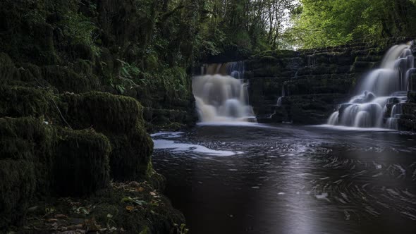 Time lapse of dark spring forest cascade waterfall surrounded by trees with rocks in the foreground alt