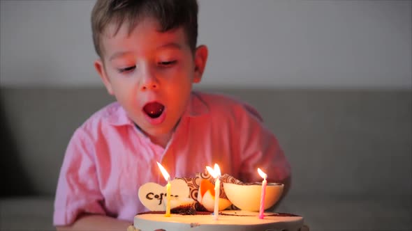 Happy Little Boy of Four Sits on the Couch Looking at the Burning Candles on the Birthday Cake