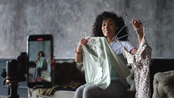 AfricanAmerican Fashion Blogger Recording Video with a Smartphone at Home Choosing Clothes Sitting