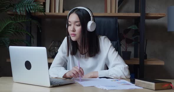 Focused Young Asian Student Wearing Wireless Headphones Studying on Online Courses Using Paper Notes