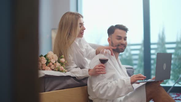 Handheld Man Freelancer Sitting on the Floor in a Bedroom and Working on a Laptop His Girlfriend is