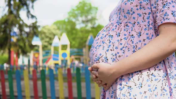 A Pregnant Woman in a Summer Dress with a Floral Print Stands on the Background of a Playground