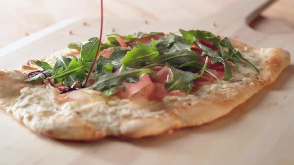 Camera follows pouring balsamic oil on a white pizza with prosciutto and arugula. Slow Motion.