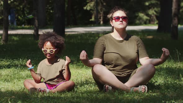 White Woman and Her Black Daughter in Sunglasses Sitting on the Grass in the Park and Meditating