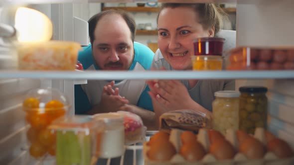 View From Fridge of Happy Fat Young Couple Taking Fatty Food From Refrigerator