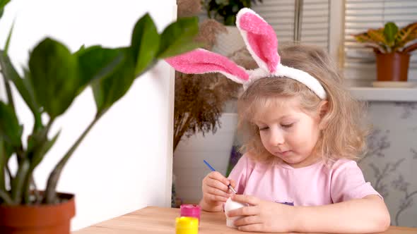 a Little Blonde Girl is Decorating an Easter Egg and Smiling at Home at the Table