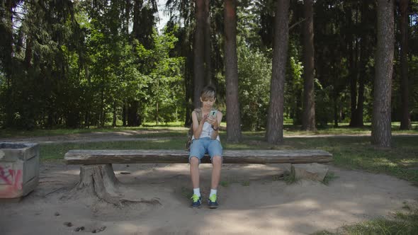 Funny Boy with a Backpack Sits on a Bench in the Forest and Uses the Phone