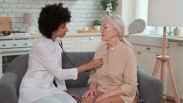 Afro American Woman Doctor Listens to Breath of Senior Woman Using Stethoscope While Sitting on Sofa