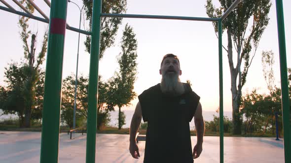 Fit Handsome Middle Aged Man with Long Gray Beard Doing Pullups on Horizon Bar on Sports Ground with