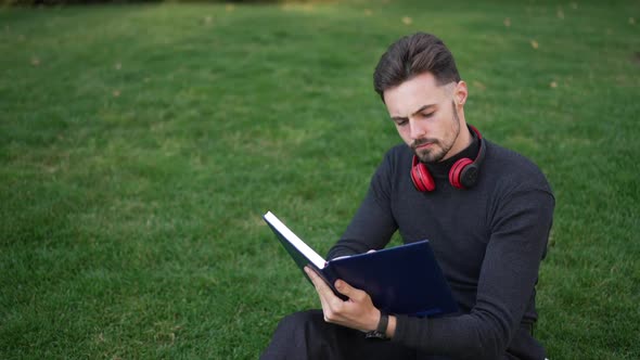 High Angle View of Concentrated Young Man Reading Checking Plans Sitting on Park Meadow Outdoors