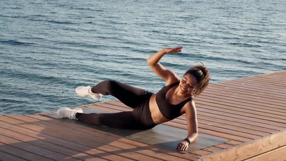 Young Sporty African American Lady Athlete Exercising on Wooden Pier Near Sea Practicing Legs