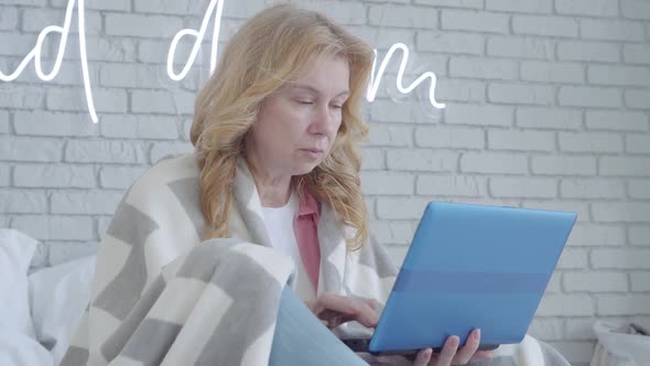 Portrait of Mid-adult Blond Woman Typing on Laptop Keyboard and Coughing. Ill Senior Caucasian