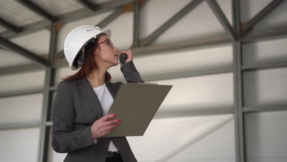 A Young Woman in a Helmet with Documents Speaks on a Walkie-talkie at a Construction Site. The Boss