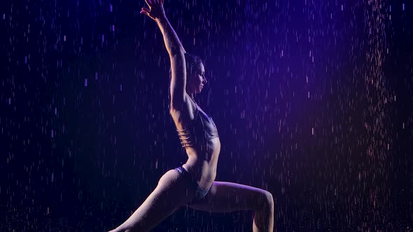 Young Woman Performs Yoga Exercises with Studio Blue Light and Water Rain. Female Body in Drops