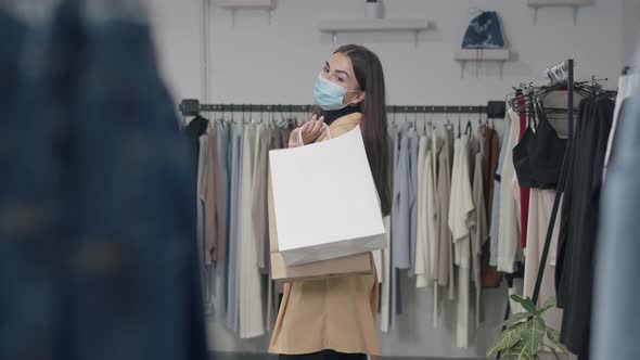 Young Confident Woman with Shopping Bags in Coronavirus Face Mask Looking Away Standing in Shop