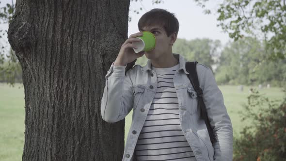 Middle Shot Portrait of Thoughtful Relaxed Young Man Drinking Coffee Standing at Tree Trunk and
