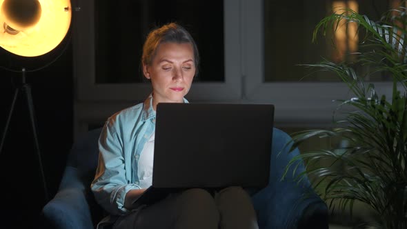 Woman is Sitting in the Armchair and Working on a Laptop at Night or Texting Someone