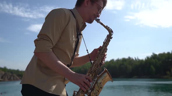 Close-up of a Man in Glasses and a Shirt Playing a Saxophone on a Sunny Day on the River Bank