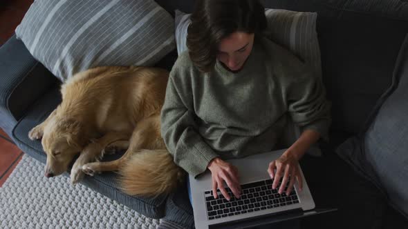 Smiling caucasian woman using laptop working from home with her pet dog on sofa next to her