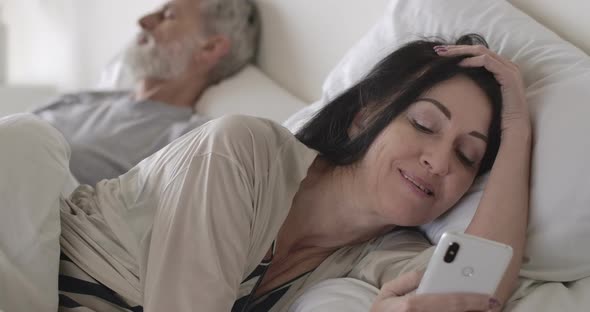 Close-up Portrait of Senior Brunette Caucasian Woman Using Smartphone Lying in Bed As Husband