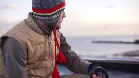 Young Caucasian Male Sitting Near Sea Lost in Thought Wearing Watch Cap and Beige Jacket Looking at