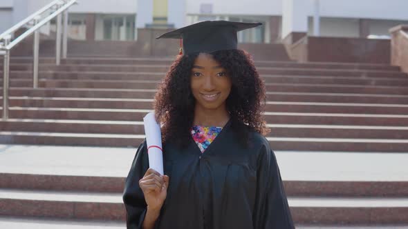 Young African American Female Graduate Standing in Front of the Camera with a Diploma and Books in