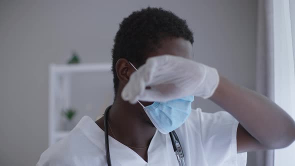 Tired African American Doctor in Face Mask Rubbing Forehead with Hand Looking Out the Window in